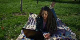时髦女孩躺在草坪上用笔记本电脑打字