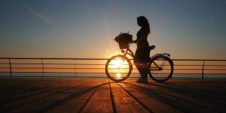 年轻女子的剪影穿着复古自行车和鲜花走在海边的木堤岸在日出或日落。浪漫的旅行概念。美丽的景色