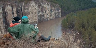 后视图一对徒步旅行者坐在山河上的悬崖边上拥抱