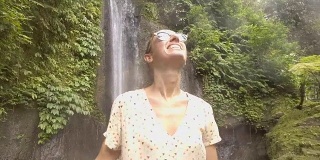 慢动作年轻女子享受新鲜瀑布在巴厘岛，印度尼西亚，亚洲。人们旅游乐趣自然的概念。女孩伸着胳膊