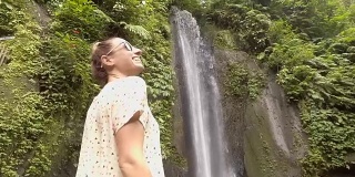 年轻女子在巴厘岛的瀑布上凝视大自然，享受大自然的新鲜。人们出行绿色环保理念。慢动作拍摄
