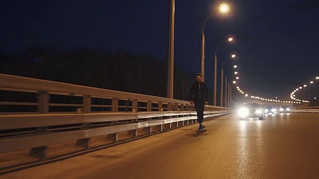那家伙晚上骑着长板穿过城市。年轻人晚上在马路上滑冰。慢动作