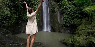 慢动作年轻女子享受新鲜瀑布在巴厘岛，印度尼西亚，亚洲。人们旅游乐趣自然的概念。女孩伸着胳膊