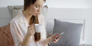 在假期的早上，当一个黑发女孩醒来的时候，她在床上喝着咖啡，在她的智能手机上打字