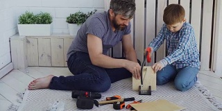 专心的孩子在用手锯锯胶合板，他的父亲用手拿着木头，帮助他的儿子教他。分享经验的概念。