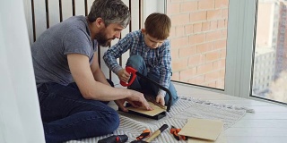 在父亲的帮助和教导下，小男孩专注于用手锯锯一块木头。团结家庭，建设工作和童年理念。