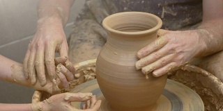 两只手交织在一起，在陶工转盘上形成一个陶罐。一个男人的手帮助一个女人的手塑造一个粘土形状。
