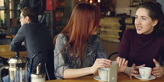 两个漂亮的女人穿着休闲衣服，在咖啡馆里聊着天，拿着咖啡杯和智能手机。很好的咖啡馆内部，顾客和家具都可以看到。