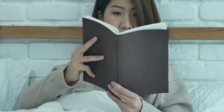 美丽的亚洲女人享受喝温暖的咖啡和看书在床上在她的卧室。亚洲女性穿着舒适的毛衣捧着一本书和一杯咖啡。生活方式亚洲女性在家的概念