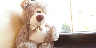 泰迪熊坐在窗台上，可爱的泰迪熊靠近窗户，在她的脖子上有一只蝴蝶，浅棕色的，儿童的软玩具，一个泰迪熊在她的脖子上有一只蝴蝶