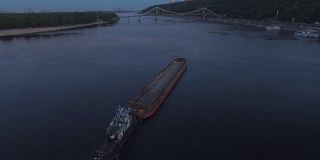 日落航拍画面显示，驳船在城市港口附近的河上航行