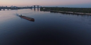日落航拍画面显示，驳船在城市港口附近的河上航行