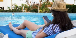 年轻漂亮的黑发女子戴着帽子躺在游泳池旁的躺椅上使用手机。3840 x2160 FullHD
