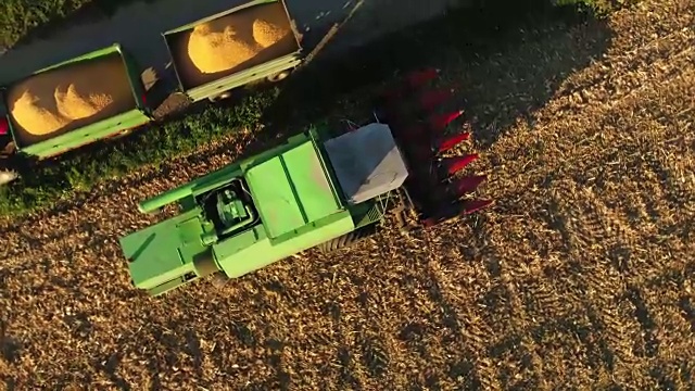 无人机视角联合收割机斜槽填充拖车收获的玉米在阳光明媚的农村田野，慢镜头