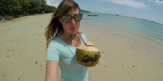 自拍:微笑的女游客漫步在沙滩上，用椰子喝饮料。