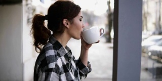 漂亮的女孩站在窗边看风景。用白色的杯子喝茶。侧视图。模糊的背景