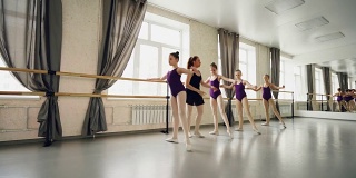 芭蕾舞学校的年轻学生正在练习基本姿势，而他们专心的女老师正在帮助他们采取正确的姿势和伸展双腿。