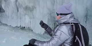 女人在贝加尔湖冰面上的旅行。冬岛之旅。女孩在冰岩石下行走。游客看着美丽的冰洞。极限跋涉和步行。背包客在大自然中休憩。