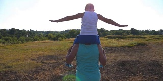父亲和儿子在阳光灿烂的夏日操场上玩耍。小男孩坐在爸爸的肩膀上，举着手玩飞机。父母和孩子一起玩。慢镜头后景特写