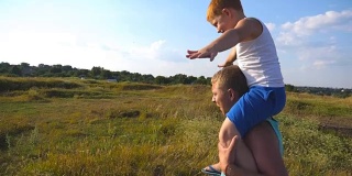 小男孩坐在爸爸的肩膀上，举着手玩飞机。阳光明媚的夏日，快乐的父子在操场上玩耍。爸爸和孩子在大自然里玩得很开心。慢动作特写侧视图