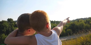 小男孩和他的父亲坐在山上，指着什么东西。父亲和他的小儿子一起花时间在户外日落。特写慢镜头后视图