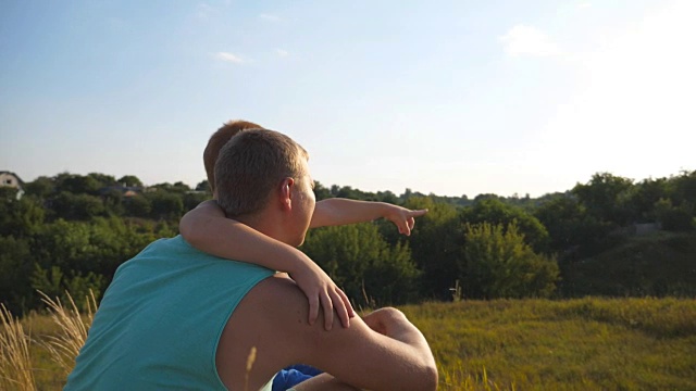 一个孩子坐在山上，指着什么东西指着他的爸爸。父亲和他的小儿子花时间一起在户外日落。慢镜头后景特写
