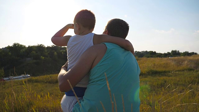 父亲和他的小儿子花时间一起在户外日落。爸爸和孩子坐在山上的绿草地上，看着周围的大自然。慢镜头后景特写