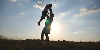 快乐爸爸在草地上吐他儿子的剪影。日落时分，父亲和儿子在操场上玩耍。爸爸和孩子在一起玩。慢动作侧视图特写