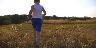 在阳光明媚的一天，一个小男孩在草地上跑步。孩子在草坪上慢跑，太阳耀斑在背后。一个男孩在夏日草地上走进大自然。慢镜头后视图