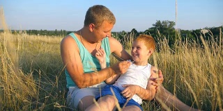 父亲在室外给小儿子挠痒痒。一个快乐的年轻人坐在草地上和他的宝贝儿子玩。快乐的爸爸和他的孩子一起在草地上玩。慢镜头