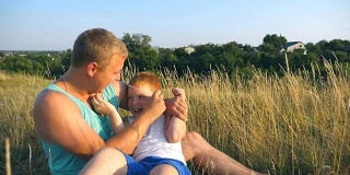 一个快乐的年轻人坐在草地上和他的宝贝儿子玩。父亲在室外给小儿子挠痒痒。快乐的爸爸和他的孩子一起在草地上玩。慢镜头