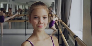 特写的美丽的小女孩在紧身衣站在芭蕾舞班，微笑着看着相机。其他学生在后台做练习。