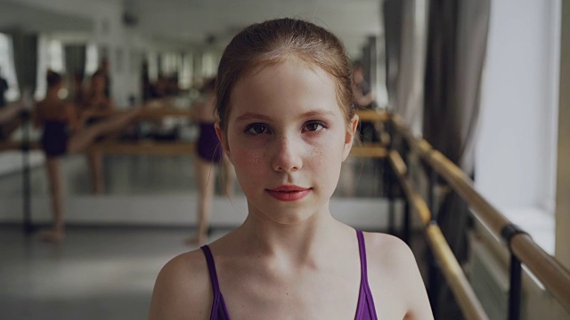 在宽敞的灯光舞厅里，化妆的年轻女孩在上芭蕾课时看着镜头微笑。艺术和童年的概念。