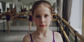 在宽敞的灯光舞厅里，化妆的年轻女孩在上芭蕾课时看着镜头微笑。艺术和童年的概念。