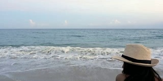 后视图幸福的女人在帽子坐在沙滩椅上热带海滩和蓝色的天空背景与海浪冲击海岸。独自女性旅行者享受户外暑假。