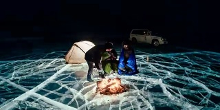 三个旅行者站在篝火旁，就在夜晚的冰面上。营地冰。帐篷紧挨着火。人们正在火炉旁取暖。延时与圆周运动。贝加尔湖。