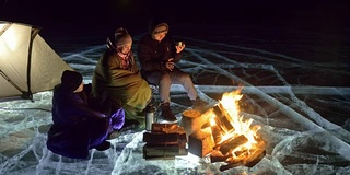 三个旅行者在夜间的冰面上着火。营地在冰上。帐篷紧挨着火。贝加尔湖。附近有一辆车。人们围着篝火取暖，穿着睡袋。