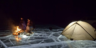 三个旅行者在夜间的冰面上着火。营地在冰上。帐篷紧挨着火。贝加尔湖。附近有一辆车。人们围着篝火取暖，穿着睡袋。