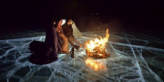 三个旅行者在夜间的冰面上着火。营地在冰上。帐篷紧挨着火。贝加尔湖。附近有一辆车。人们围着篝火取暖，穿着睡袋。