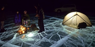 四个旅行者在夜间的冰面上着火。营地在冰上。帐篷紧挨着火。人们围着营火取暖。摄影师在用三脚架拍摄相机。
