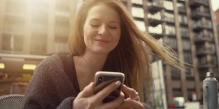 漂亮的红头发女人坐在长凳上微笑着使用智能手机，连接女孩写信息。华丽的纽约城市景观的背景。