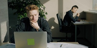 年轻人在书桌前用笔记本电脑工作，同事在现代办公室写作