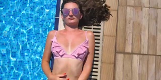 近距离的年轻女子在粉红色比基尼躺在盆的边缘和直她的太阳镜。美丽的女孩在酒店游泳池附近晒太阳。暑假或假期的概念。俯视图