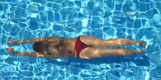 俯视图，不认识的年轻女子游泳在游泳池的水和出现在阳光明媚的一天的表面。美丽的女孩穿着红色比基尼漂浮在盆地的酒店。假期或假日概念。