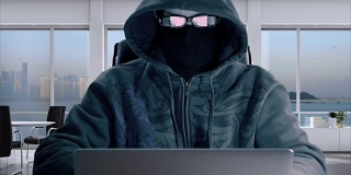 匿名程序员在掩码编码和打字在笔记本电脑在办公室。戴着眼镜戴着巴拉克拉瓦帽的黑客正在输入程序代码。