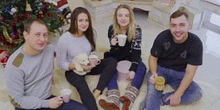 四个年轻人坐在圣诞树附近的地板上喝茶或咖啡