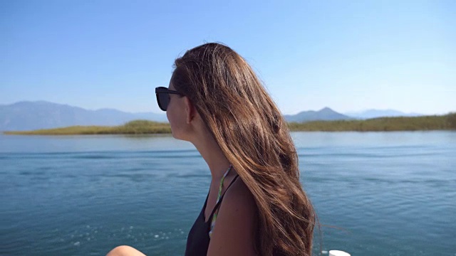 小女孩坐在船头，欣赏沿途美丽的自然景观。快乐的女人享受夏天的旅行。假期或假日概念。特写慢动作