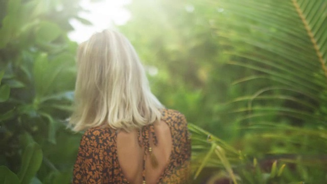 一个美丽的年轻金发女人穿着裙子走过热带岛屿上郁郁葱葱的森林的背影拍摄。