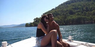 在阳光明媚的日子里，一对年轻的情侣坐在船头，对着摄影师摆姿势。幸福的情侣在船上共度时光，享受夏天的旅行。假期的概念。近距离