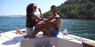 在阳光明媚的日子里，一对年轻的情侣坐在船头，对着摄影师摆姿势。幸福的情侣在船上共度时光，享受夏天的旅行。假期的概念。近距离